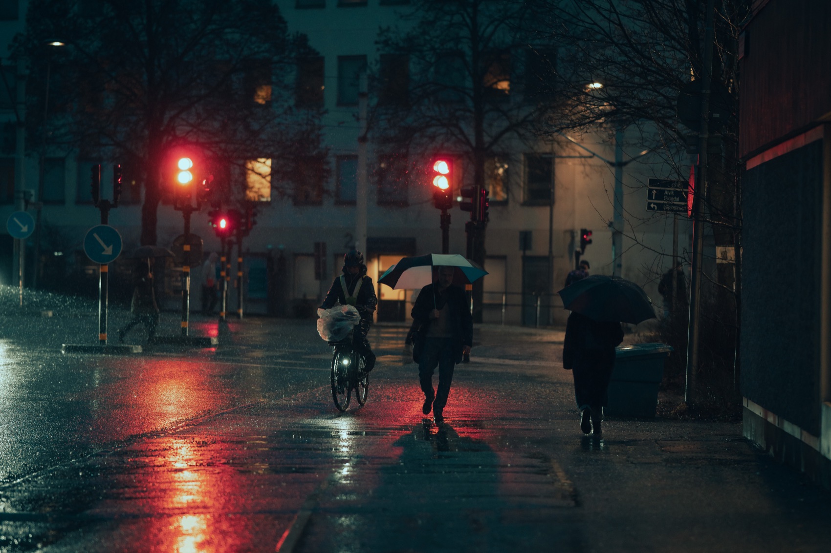 Umbrella-carrying pedestrians crossing a rain-soaked street under red lights at blue hour