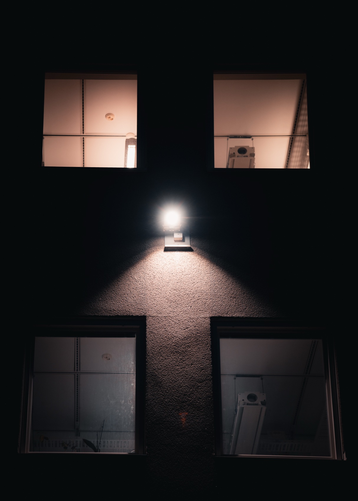 Four warm-lit building windows in deep darkness, with one central exterior lamp casting a triangular pool of light