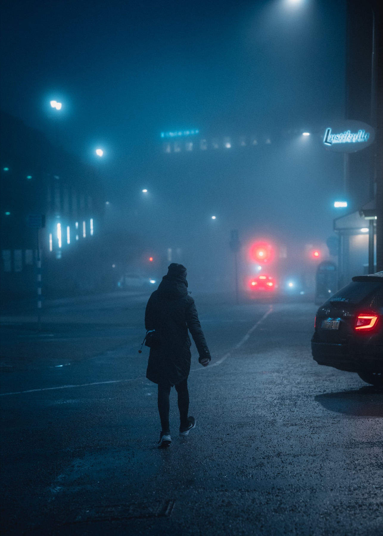A solitary pedestrian on a fog-filled blue night street with distant red lights