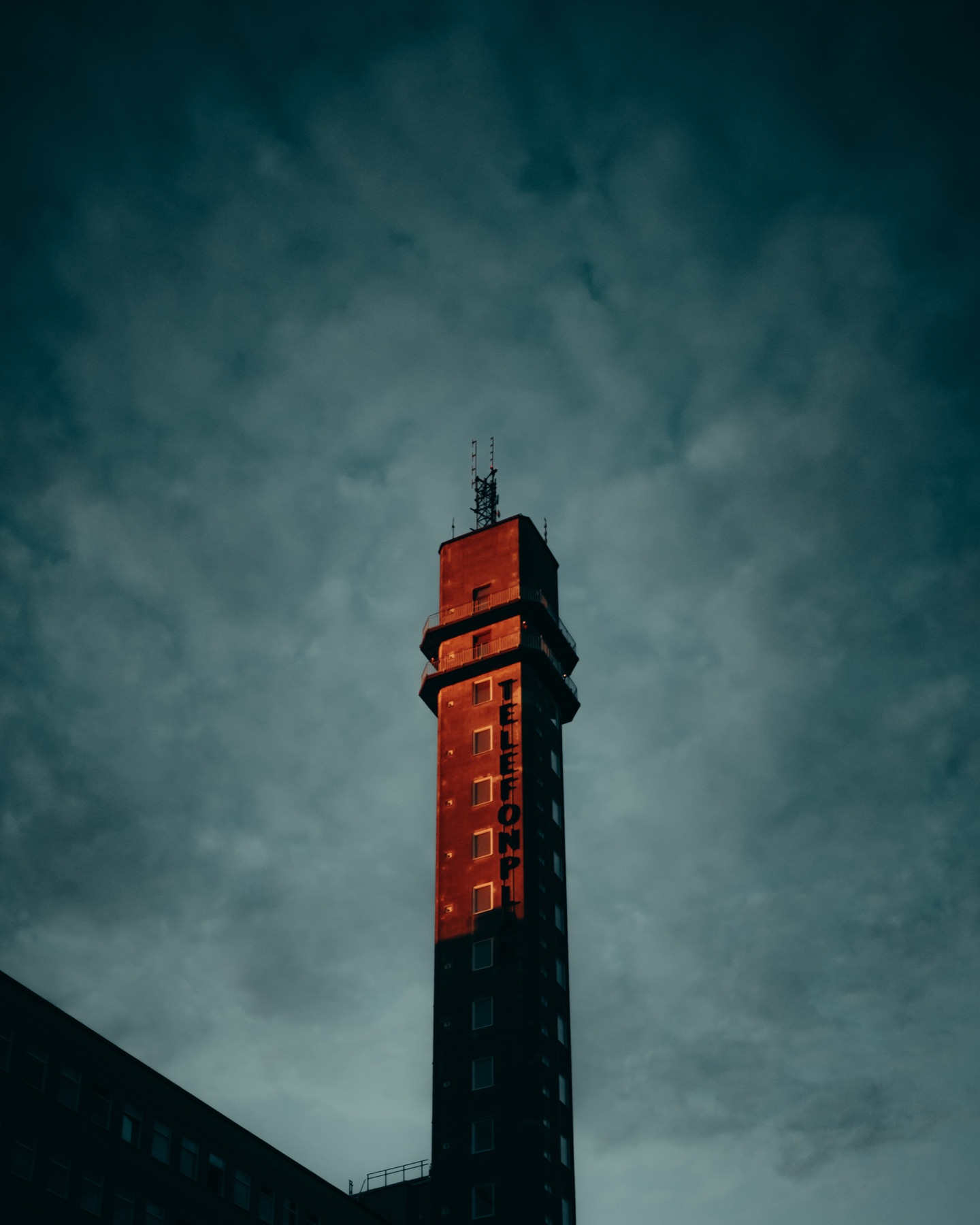 A red-orange illuminated tower under a dramatic stormy blue sky